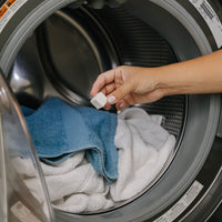 Hand placing a laundry detergent cube into a washing machine with clothes inside