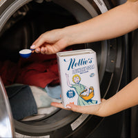 A person holding a tin of Nellie's Laundry Soda and a blue measuring scoop in front of an open front-loading washing machine, with laundry visible inside.