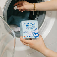 A person holds a box of Nellie's Laundry Cubes in front of an open front-loading washing machine, with a hand dropping a cube into the drum.
