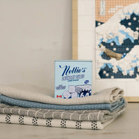 A stack of folded patterned towels in neutral and blue tones sits in front of a framed art print, with a small box labeled Nellie's Laundry Cubes standing upright in the center.