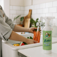 A bottle of Nettie's Fruit & Veggie Wash stands on a white kitchen counter in a bright kitchen, with a person washing vegetables in the sink in the background.