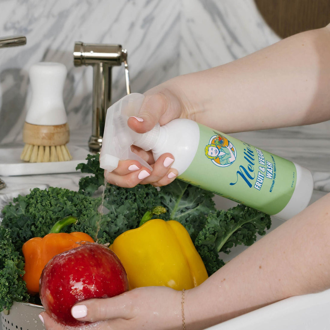 A person's hands washing fresh vegetables, including a red apple, yellow and orange bell peppers, and kale, under running water with Nellie's Fruit & Veggie wash.