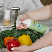 A person's hands washing fresh vegetables, including a red apple, yellow and orange bell peppers, and kale, under running water with Nellie's Fruit & Veggie wash.
