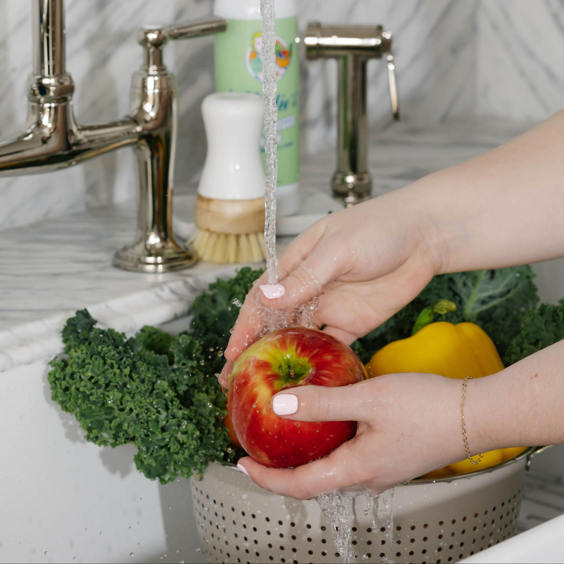 Person washing an apple under a faucet with a marble backsplash.