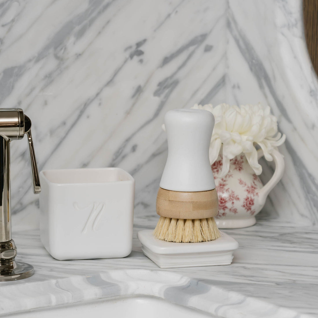 Bathroom sink area with Nellie's Kitchen Brush, Nellie's Dish Butter, and decorative items on a marble countertop.