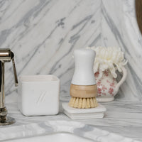 Bathroom sink area with Nellie's Kitchen Brush, Nellie's Dish Butter, and decorative items on a marble countertop.