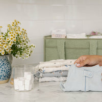 A hand holds a light blue and white striped baby romper in the foreground, with folded baby clothes stacked on a marble countertop. A blue and white floral vase with white daisies, a glass jar of Nellie's Laundry Cubes, and a sage green storage cabinet are visible in the background against a white tiled wall.