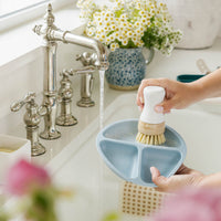 A light blue three-compartment baby feeding tray being washed with Nellie's Kitchen Brush under running water in a kitchen sink. A person's hands are visible holding the brush, with flowers and a window in the background. 