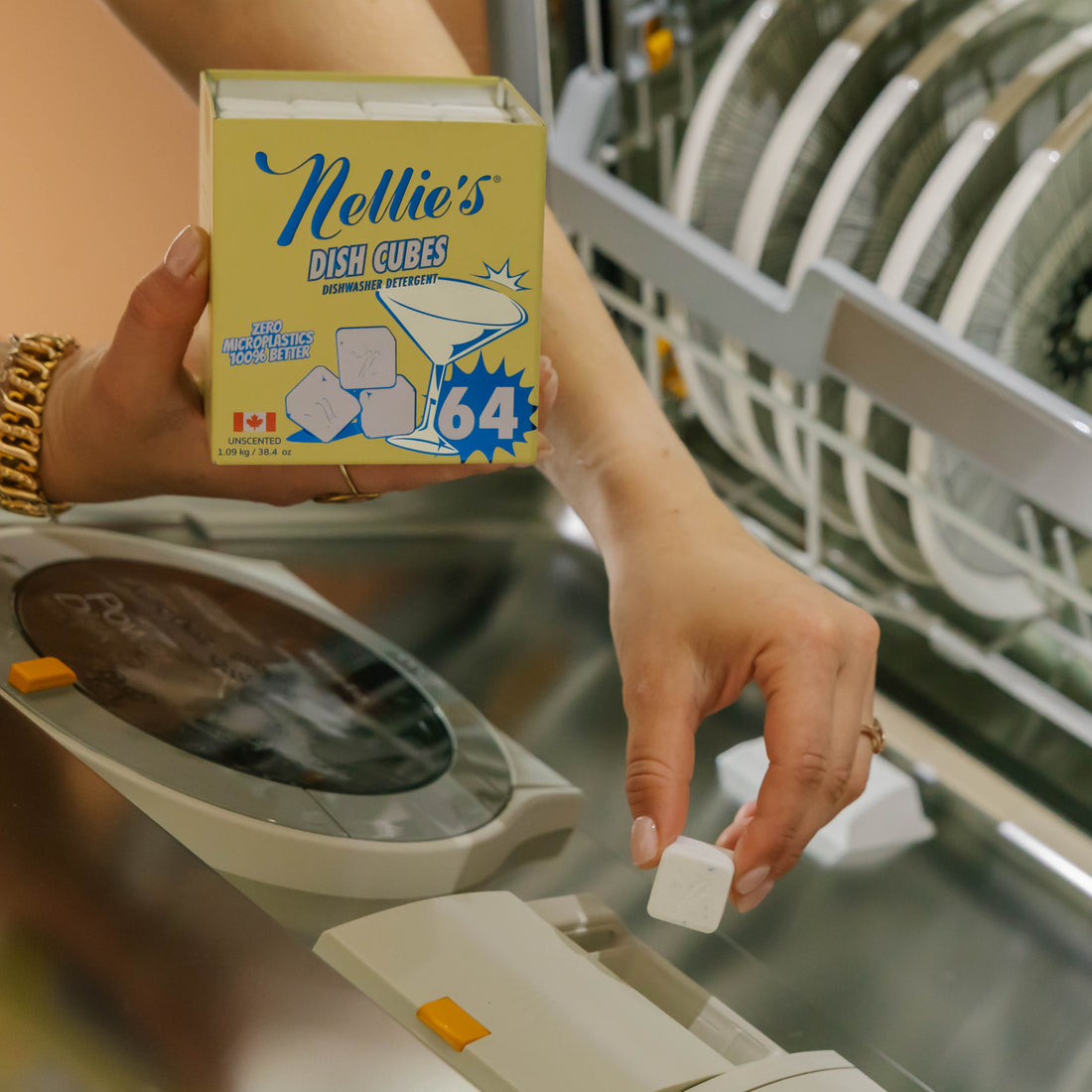 A person holds a box of Nellie's Dish Cubes in one hand and reaches to pick up a small white cube with the other hand, with a dishwasher visible in the background.