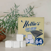 A box of Nellie's Dish Cubes with several white cubes placed in front, styled on a light surface with a blurred green plant in the background.