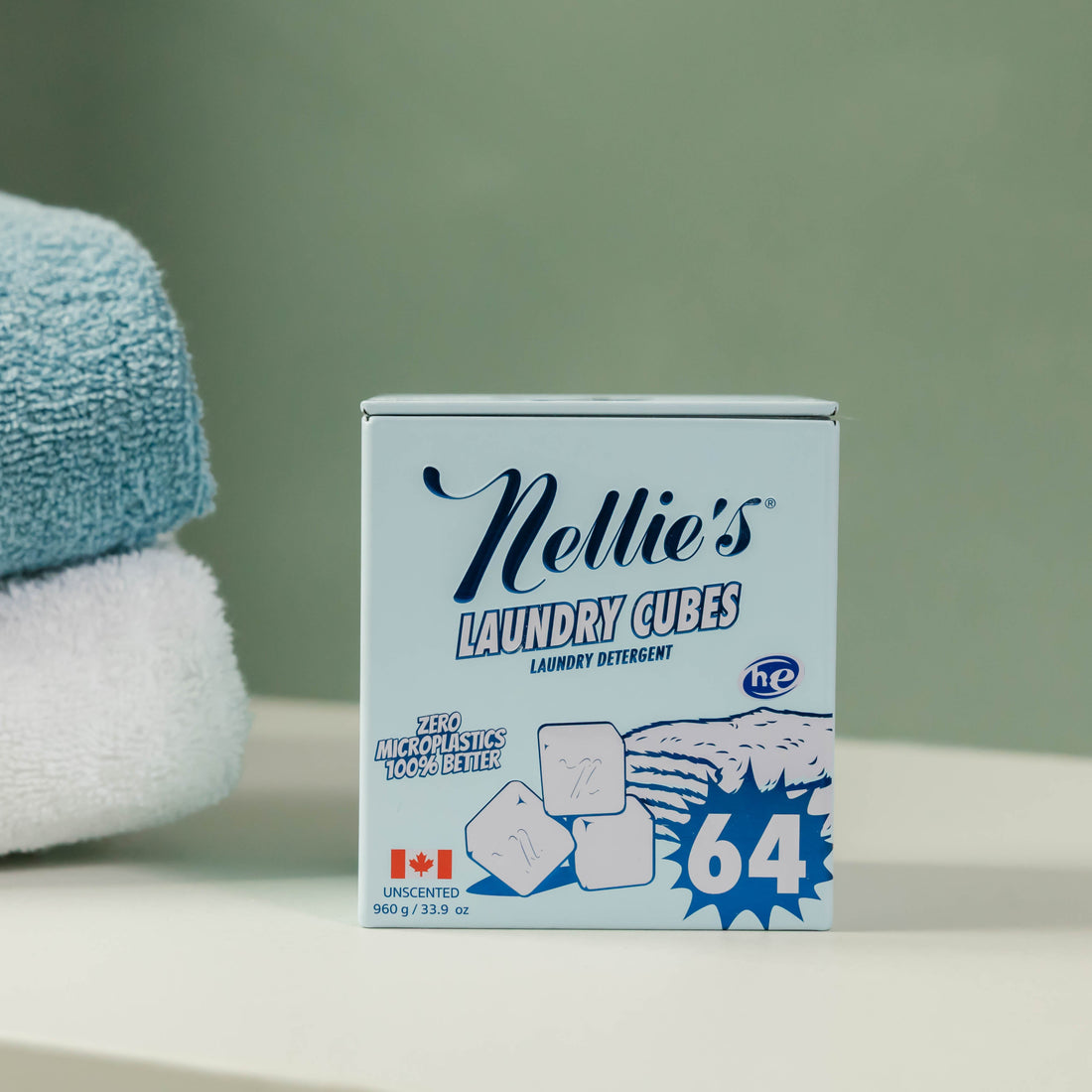 A box of Nellie's Laundry Cubes 64 unbleached laundry detergent, styled on a white surface with a soft-focus green background and folded towels on the left.