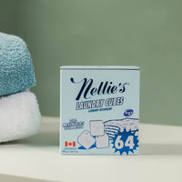 A box of Nellie's Laundry Cubes 64 unbleached laundry detergent, styled on a white surface with a soft-focus green background and folded towels on the left.